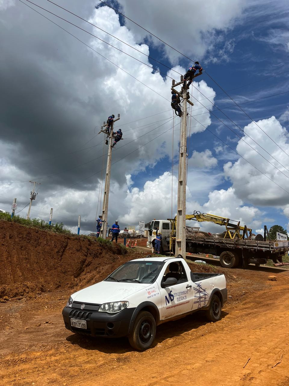 Caminhão com guindaste içando poste e equipe uniformizada da MS em deslocamento de rede de média tensão, com dois postes de concreto e cabos